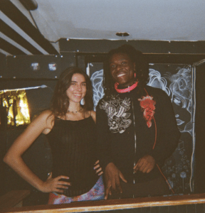 Two people stand side by side indoors, smiling at the camera. The background features a black and white mural with abstract and floral designs, adding an artistic touch to their story.